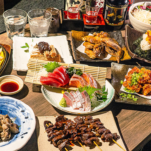 Assortment of Japanese dishes on a wooden table: sashimi platter with tuna and salmon, skewered grilled meat, fried chicken pieces, rice, and dipping sauces.