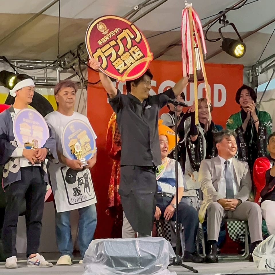 S__320569350_0_0S ‣ 名古屋の飲食店企業/有限会社 坊’S Man in black clothing raising a large red circular award sign on a stage, surrounded by people holding round signs and trophies with microphones and a seated audience in the background.