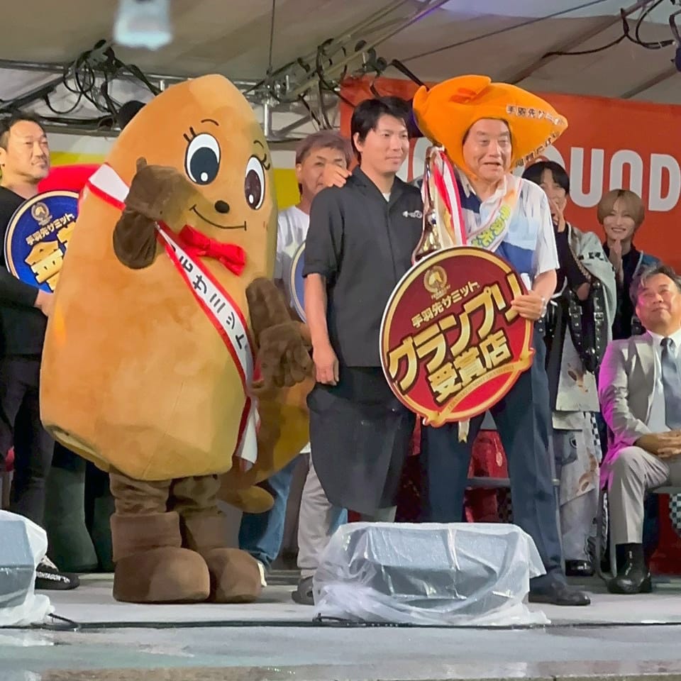 S__320569351_0_0S ‣ 名古屋の飲食店企業/有限会社 坊’S Costumed mascot in brown outfit with red bow tie posing with people on a stage during an award ceremony in Japan.