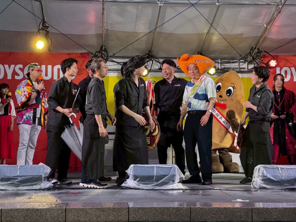 S__320569352_0_0 ‣ 名古屋の飲食店企業/有限会社 坊’S Group of performers and hosts on a stage during an event, including a man in a colorful shirt and a potato mascot in costume.