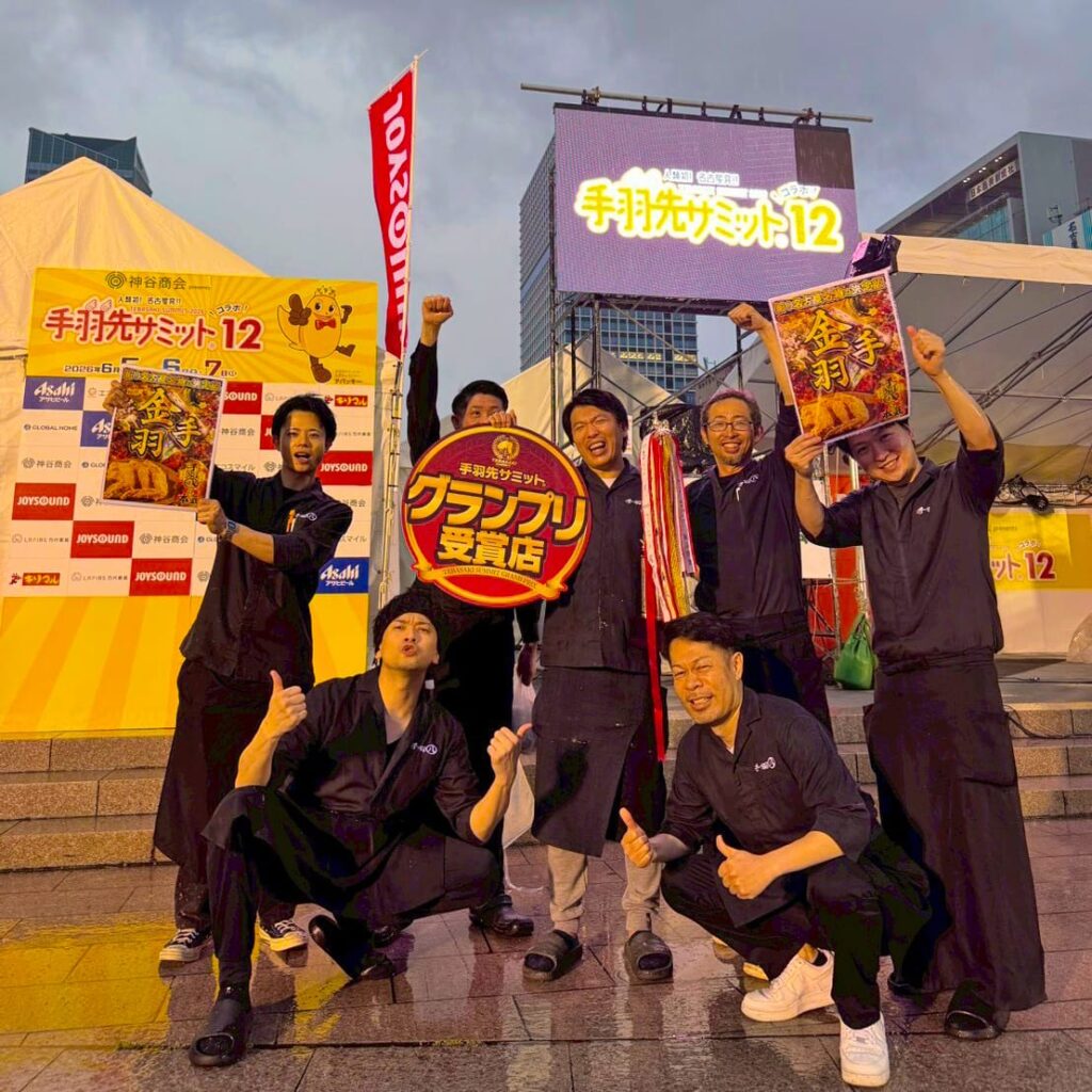 S__320569361_0S ‣ 名古屋の飲食店企業/有限会社 坊’S Seven men in black outfits pose and cheer on a stage, holding banners and a large circular award at a Japanese outdoor festival.