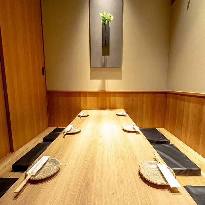 Long wooden dining table with six place settings (plates and chopsticks) and black floor cushions in a minimalist room; wall art featuring a vase centered on the beige wall.