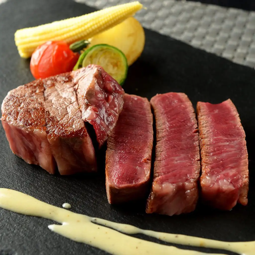 s_0nc8 ‣ 名古屋の飲食店企業/有限会社 坊’S Slices of medium-rare steak arranged on a black plate with grilled corn, tomato, and zucchini garnish and a pale sauce drizzle.