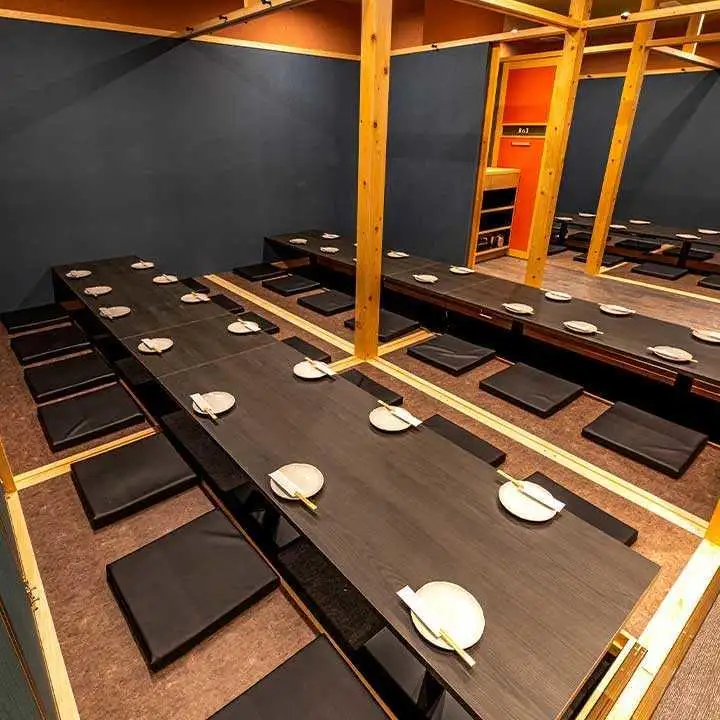 s_0nca ‣ 名古屋の飲食店企業/有限会社 坊’S Long, dark wooden tables with black cushions and white plates with chopsticks, arranged for floor seating in a wooden-framed dining room.