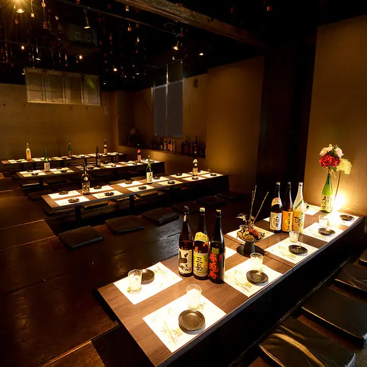 Dim izakaya with long low tables and floor seating; bottles of sake lined along the counter and warm candlelight.
