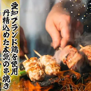 Grilled chicken skewers cooking over flames; vertical Japanese text on the left reads '愛知ブランド鶏を使用 丹精込めた本気の串焼き' (Using Aichi-brand chicken, meticulously prepared skewers).